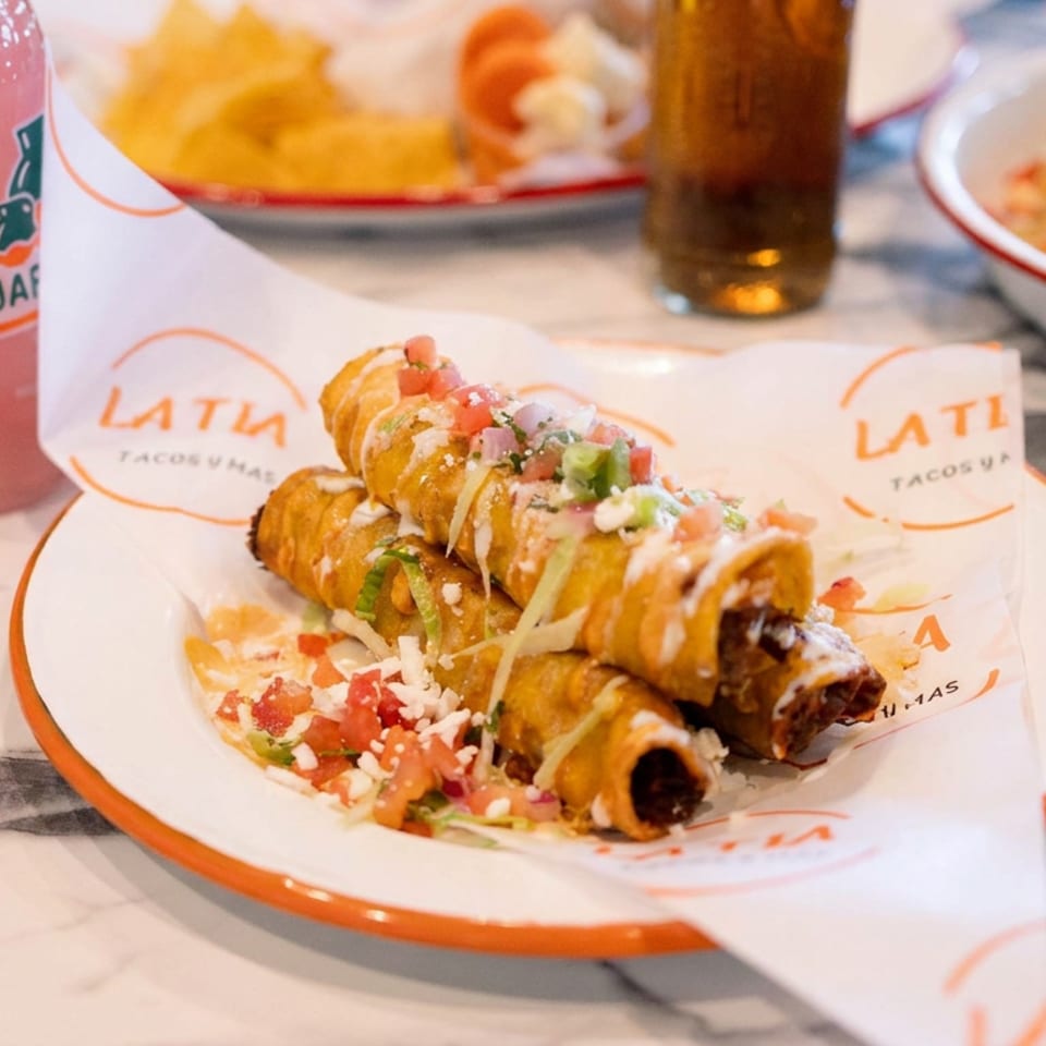 Crispy rolled tacos topped with crema, cheese, pico de gallo, and guacamole on a branded LATI Tacos plate.