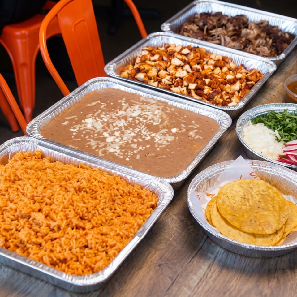 Mexican feast served in foil trays, featuring rice, beans, seasoned meat, tortillas, garnishes, and colorful sauces on a wooden table.