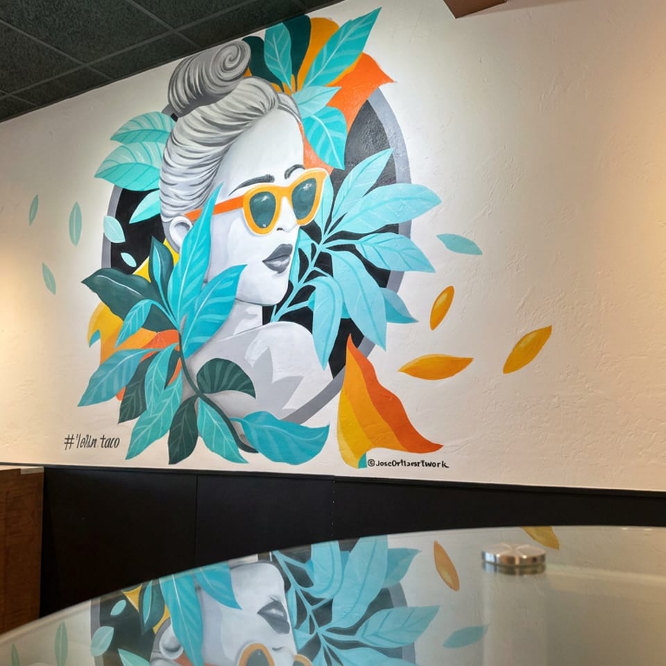 Colorful mural of a woman wearing orange sunglasses, surrounded by turquoise leaves and orange accents in a restaurant dining area.