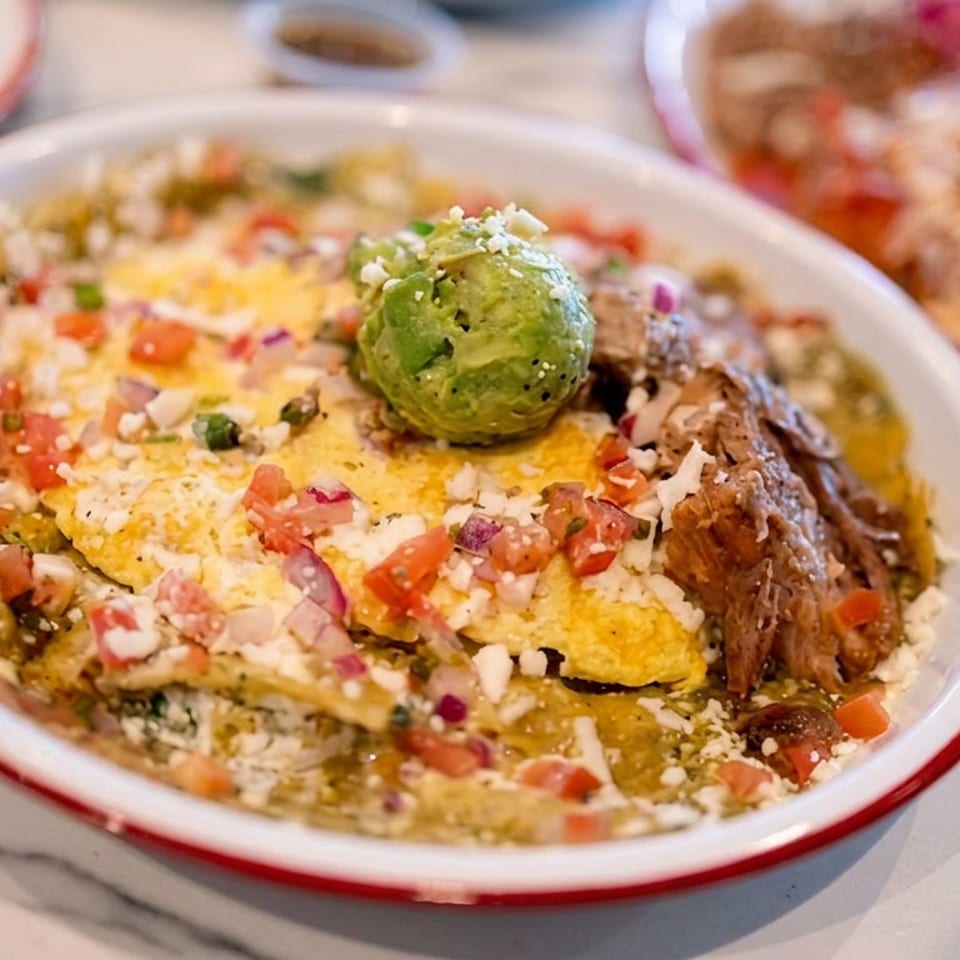 A bowl of Mexican chilaquiles topped with eggs, shredded meat, avocado, cheese, and pico de gallo on a marble table.